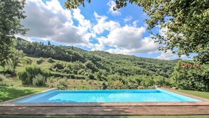 A rectangular swimming pool with a wooden deck, surrounded by well-maintained grass, set against a backdrop of rolling hills and a partly cloudy sky. - Stunning  villa for 12 guests with WIFI, private pool, TV, balcony, pets allowed and panoramic v... (Greve In Chianti)