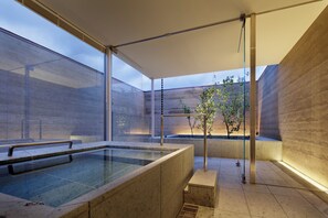 Un área de spa moderna con un jacuzzi de vidrio, un banco de madera y un pequeño árbol.