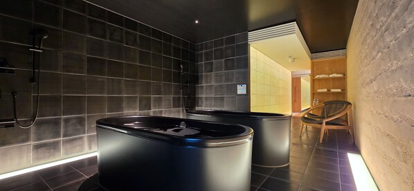 Un baño moderno con paredes de azulejos oscuros, una bañera negra y una silla de madera.