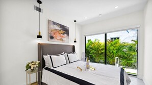 A modern bedroom with a large bed, a bedside table with a vase of flowers, a painting on the wall, and a view of greenery outside. - Calm Miami Oasis -Secluded Patio (Miami)