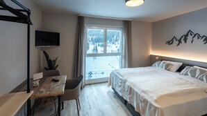 Une chambre d’hôtel avec un lit, un bureau, une chaise, une télévision et une fenêtre donnant sur un paysage enneigé.