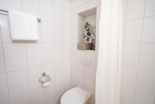 A bathroom with white tiles, a toilet, a towel rack, a shower curtain, and a shelf with a plant and a basket. - Studio 54 (Davos)