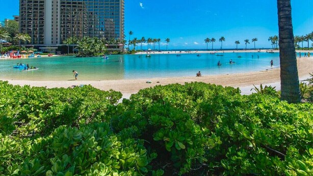 A beach with a tall hotel in the background, clear blue water, and palm trees.