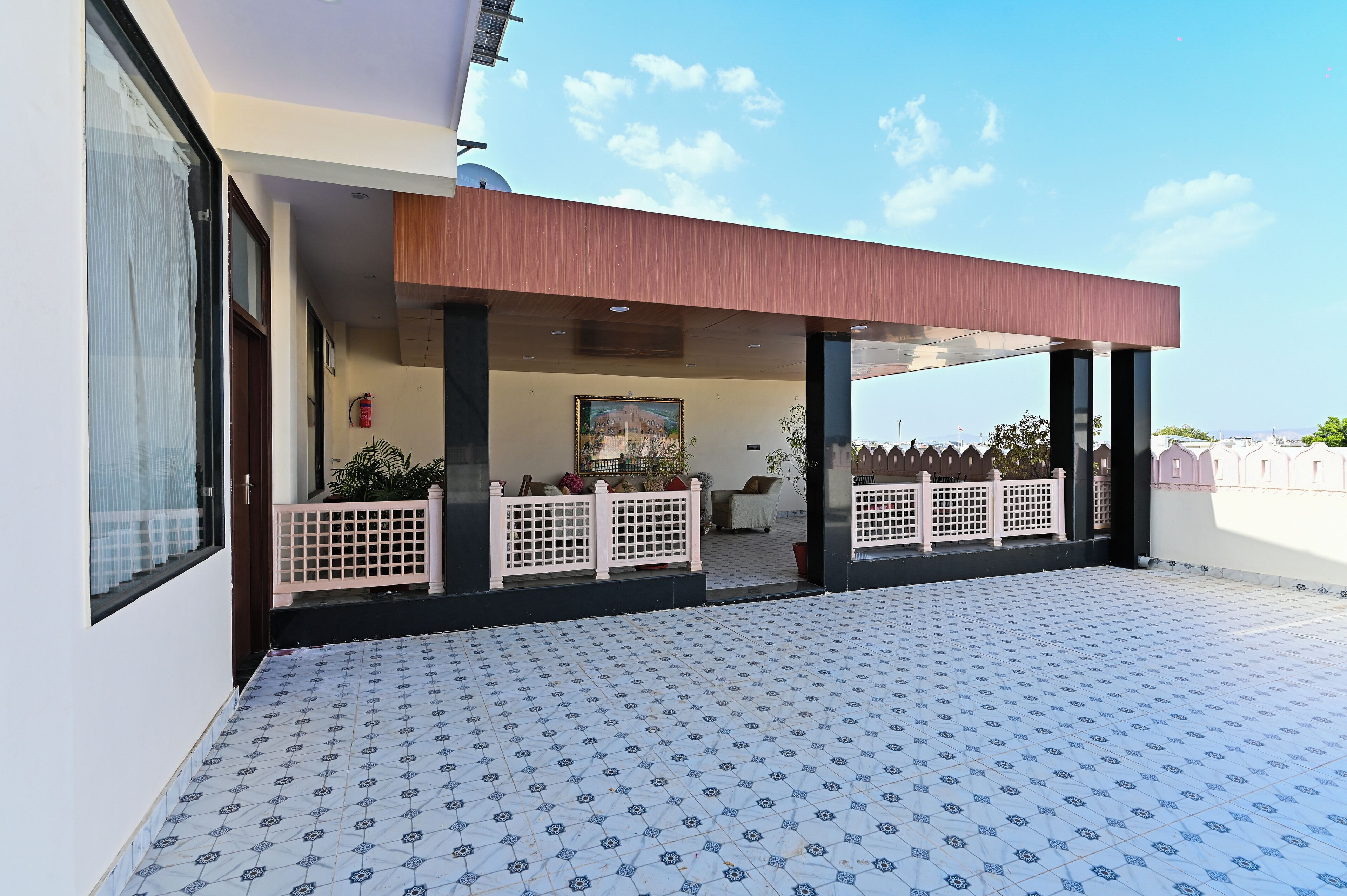 Uma área externa coberta com piso de azulejos, pérgola de madeira e vista para um edifício ao fundo.