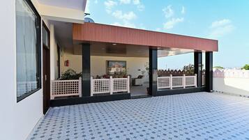 Uma área externa coberta com piso de azulejos, pérgola de madeira e vista para um edifício ao fundo.