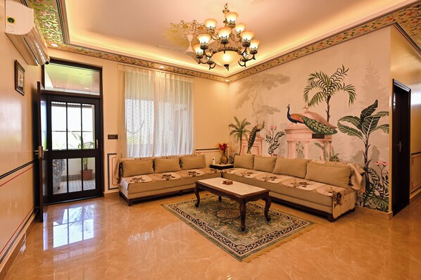 A living room with a sectional sofa, a coffee table, a chandelier, and a mural of tropical plants. - Harnawa Haveli - A Boutique Heritage Stay (Jaipur)