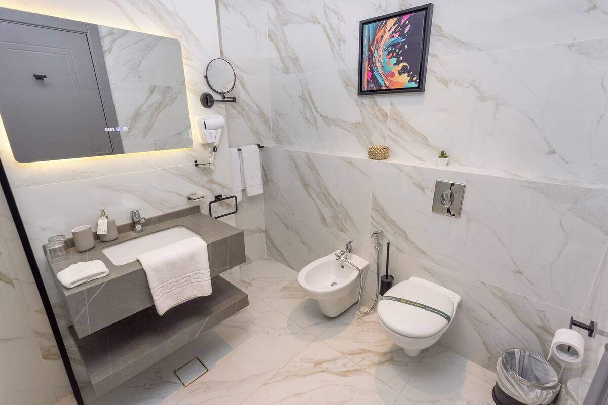 A modern bathroom with a marble floor, a large mirror, a bidet, a toilet, and a framed artwork on the wall.