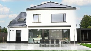 A modern two-story house with a flat roof, large windows, and a patio with a table and chairs. - Spacious, sunny luxury vacation home with sauna and garden! (Gistel)