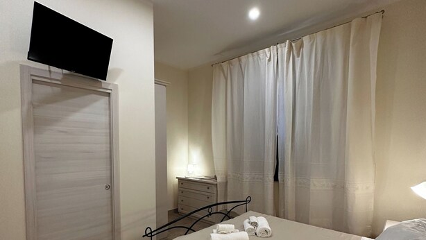 A bedroom with a bed, a bedside table, a TV mounted above a door, and a window with curtains. - Double room with private bathroom in Terralba (Oristano) - Mirto (Terralba)