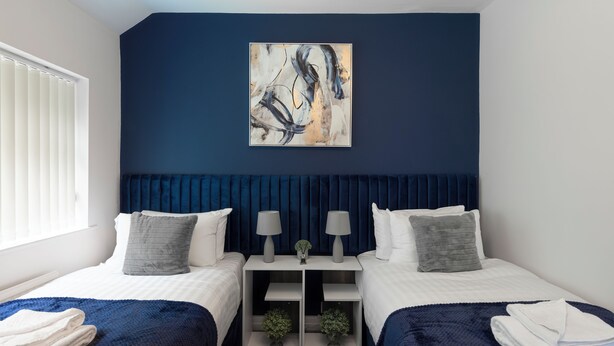 A hotel room with two beds, a dark blue accent wall, a large abstract painting, and a window with blinds.