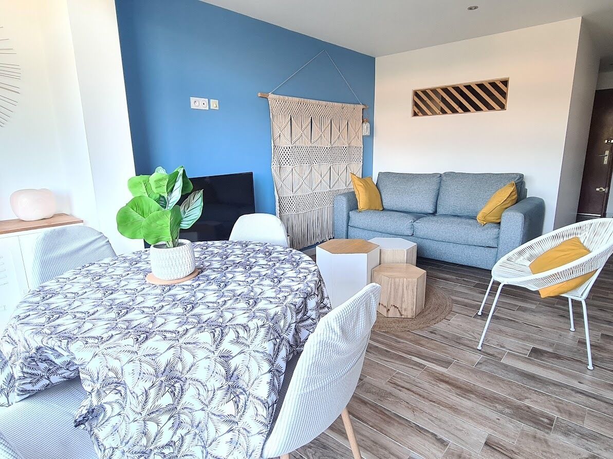 A modern living room with a blue accent wall, a grey sofa with yellow cushions, a white chair, a round dining table with a patterned tablecloth, and a large green plant.
