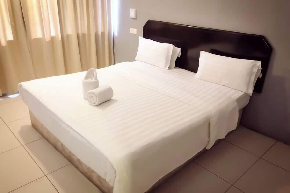 Ein ordentlich bezogenes Bett mit weißen Leinen und Kissen, ein aufgerolltes Handtuch und ein dunkles Kopfteil in einem Hotelzimmer mit Fliesenboden und beigen Vorhängen.