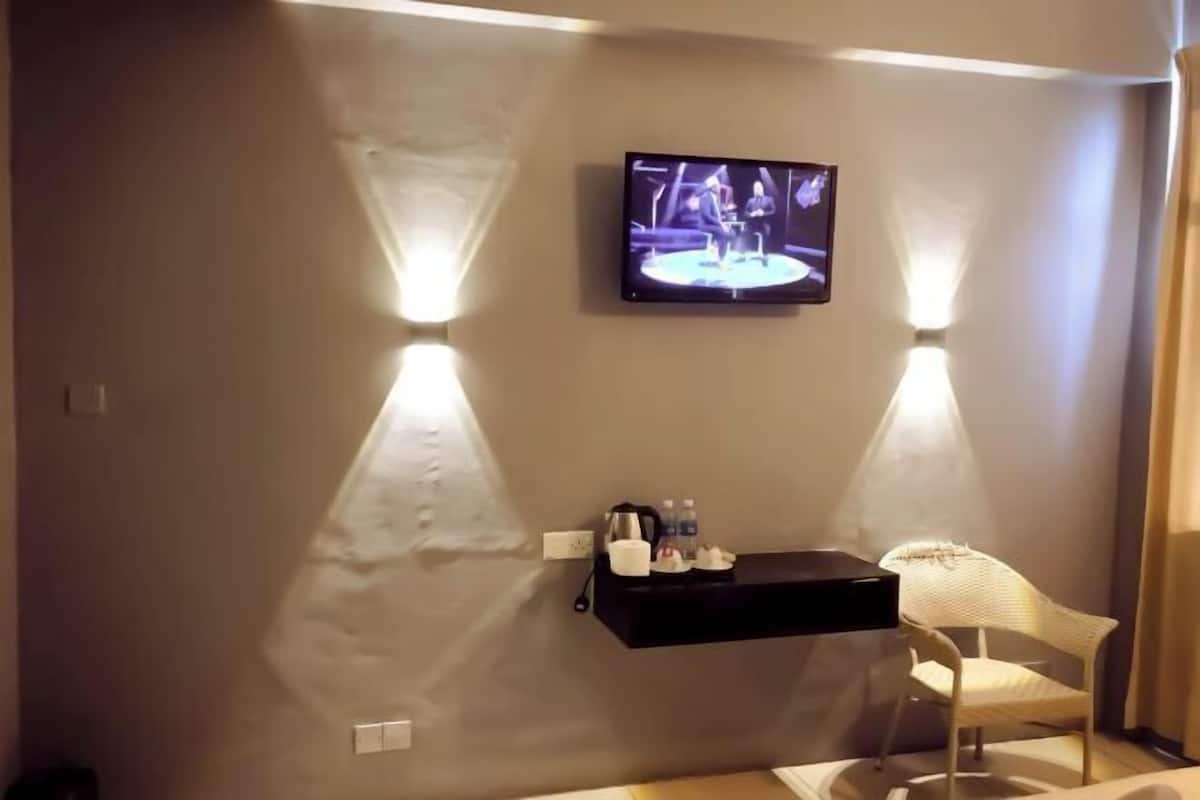 Ein Hotelzimmer mit an der Wand befestigtem Fernseher, einer kleinen Ablage für Toilettenartikel und einem weißen Stuhl.