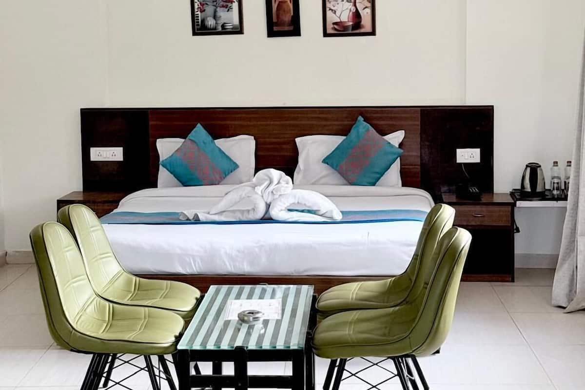 A hotel room with a bed, two green chairs, a small table, a ceiling fan, and three framed pictures on the wall.