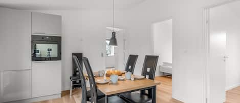 A modern kitchen with a built-in oven, a dining area with a wooden table and black chairs, and a pendant light.