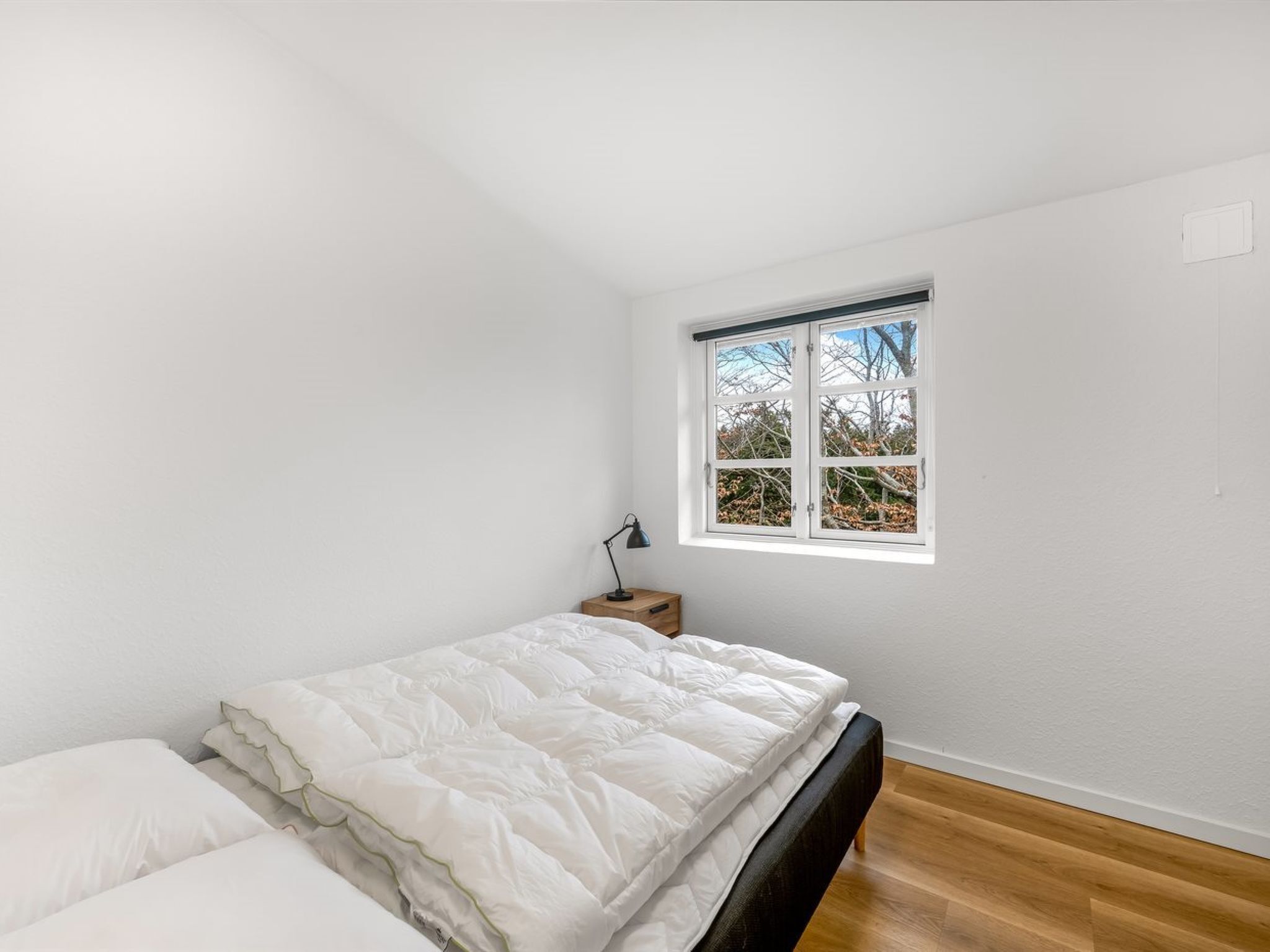 Une chambre avec un lit simple, une table de chevet, une lampe de bureau et une fenêtre donnant sur des arbres.
