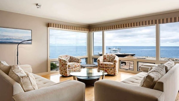 Un salon moderne doté d’une grande fenêtre donnant sur la mer, comprenant un canapé, deux fauteuils et une table basse en verre.