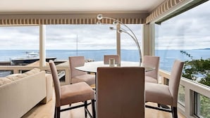 Une pièce moderne avec une table ronde et quatre chaises, de grandes fenêtres offrant une vue sur la mer et les bateaux, et un luminaire décoratif au plafond.