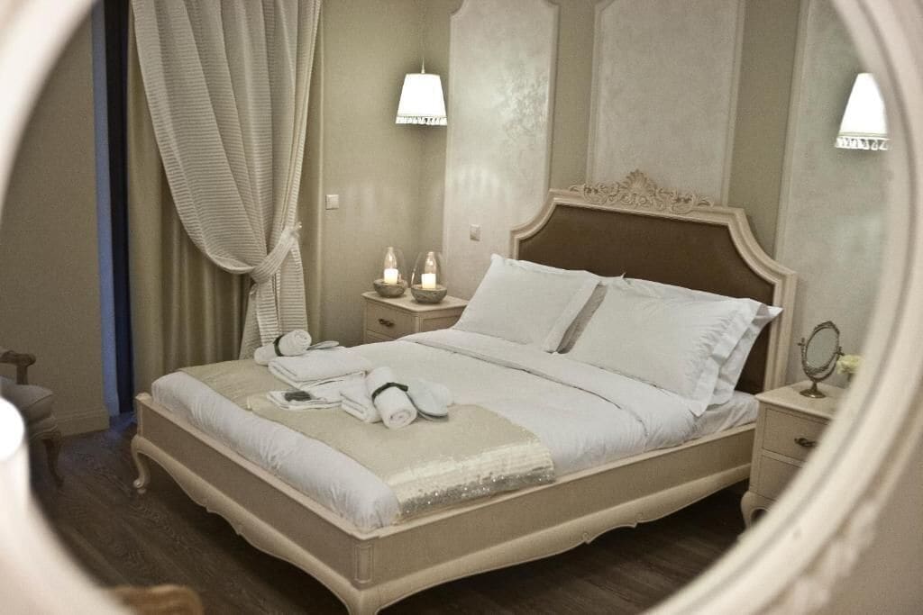 Una cama bien hecha con almohadas y una manta, una mesita de noche con lámpara, y un espejo.