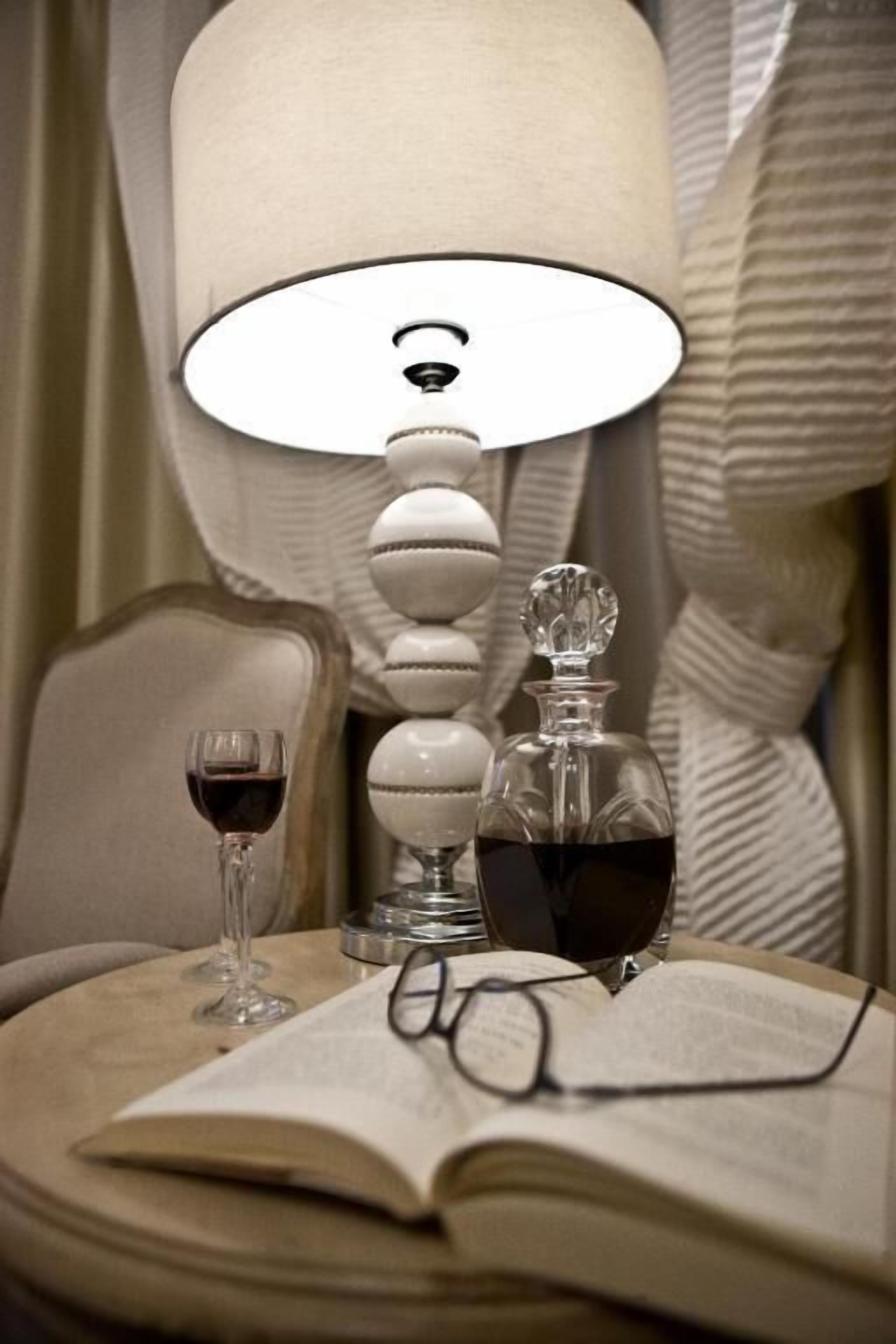 A bedside table with a lamp, a wine glass, a book, and a pair of glasses.
