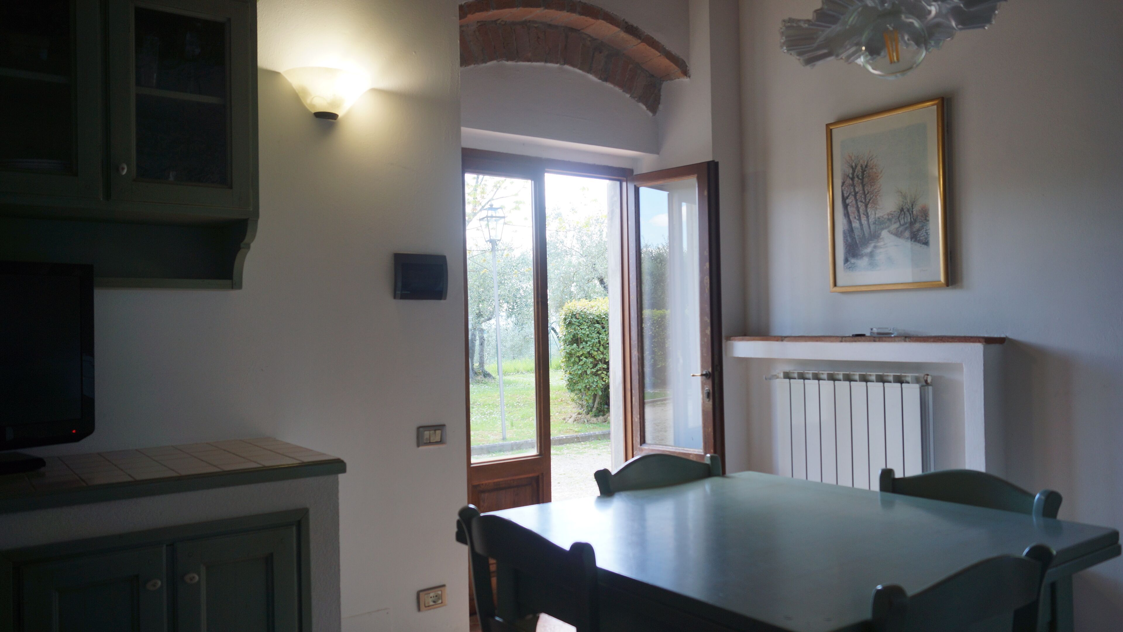Un coin repas avec une table et des chaises, un radiateur, un tableau encadré au mur et une fenêtre donnant sur un paysage verdoyant.