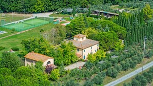 An aerial view of a residential area with a tennis court, a house with a tiled roof, and dense greenery. - Villa Panconesi (Montespertoli)