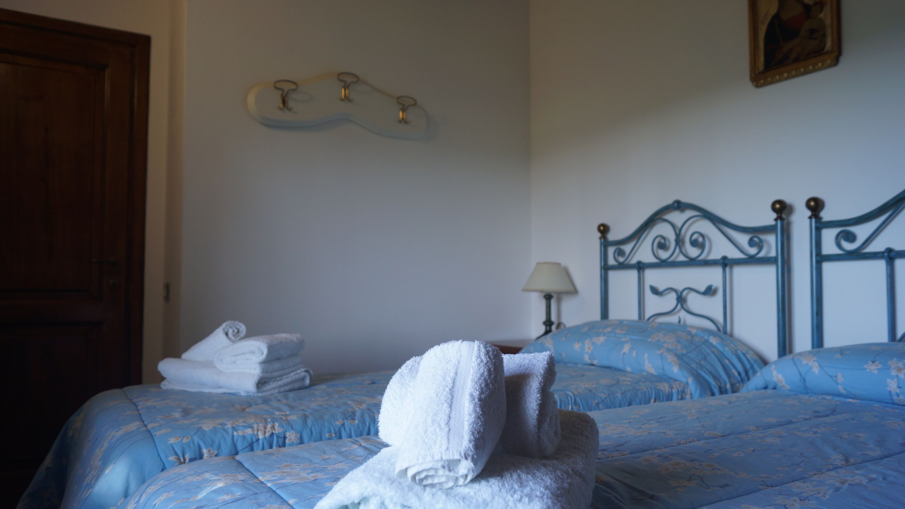 A bedroom with two beds, each with a blue floral bedspread. There are white towels on the beds, and a wooden door is visible on the left.