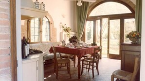 A dining area with a table set for four, wooden chairs, a brick wall, a window with curtains, and a ceiling fan. - Villa Panconesi (Montespertoli)