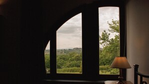 A room with a large arched window overlooking a green landscape, a wooden table with a lamp, and a chair. - Villa Panconesi (Montespertoli)