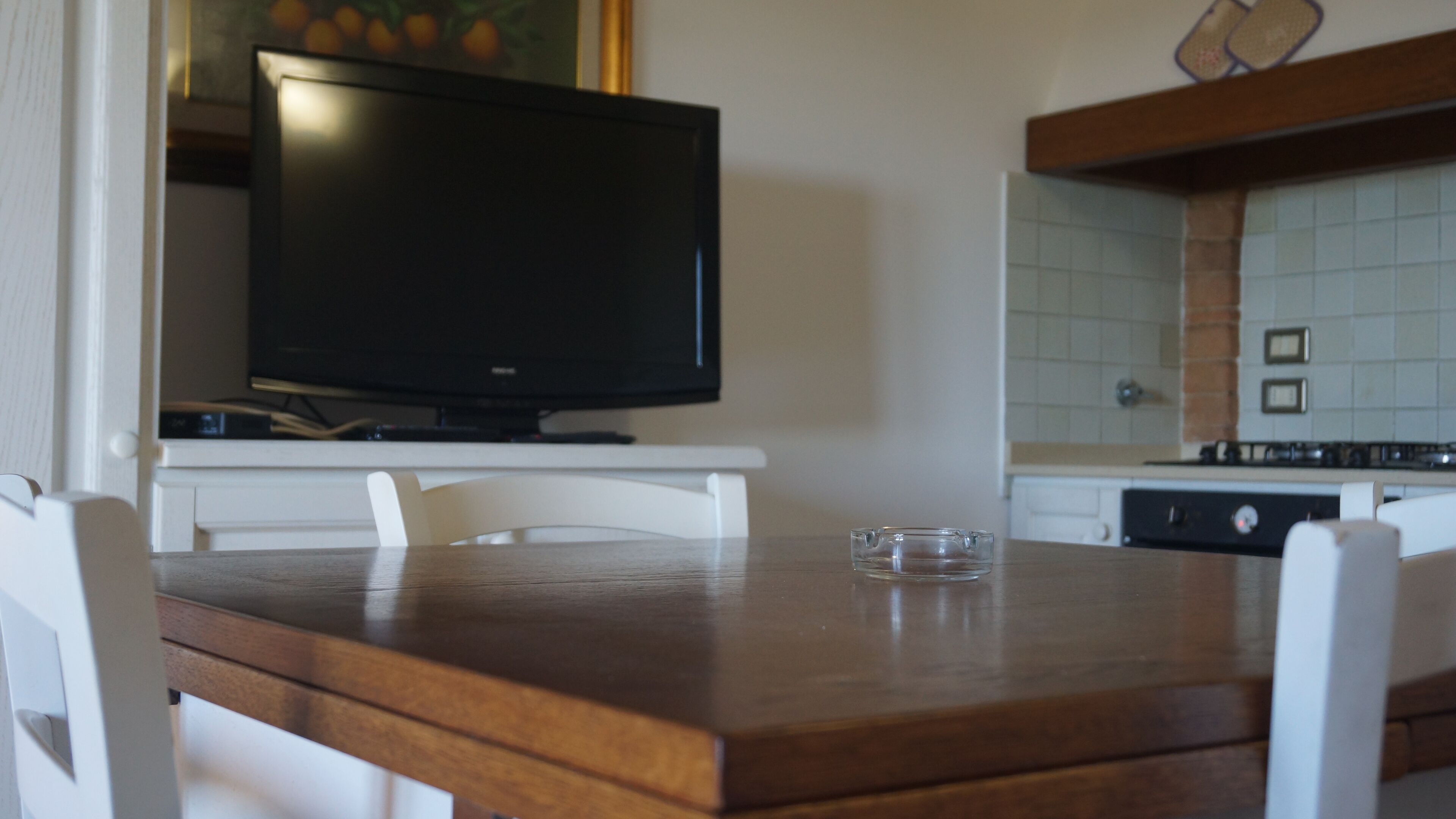 Una mesa de comedor de madera con sillas, un televisor sobre un mueble bajo y una cocina con salpicadero de azulejos y estufa.