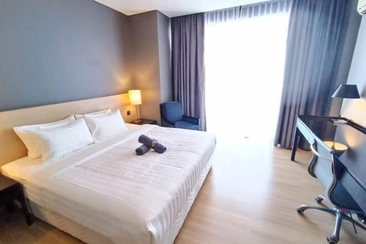 Habitación de hotel con cama, escritorio, silla, lámpara y ventana con cortinas.