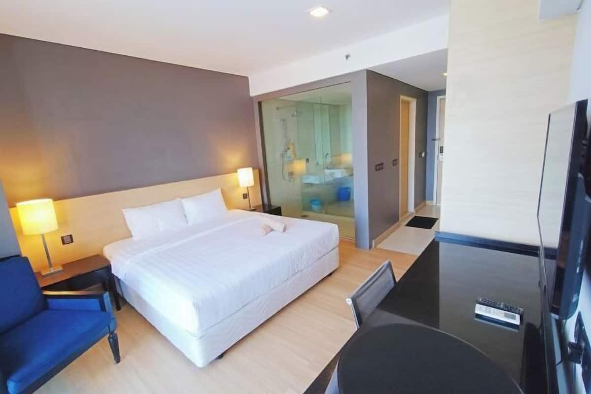 Habitación de hotel con cama, escritorio, silla, televisor y cabina de ducha de vidrio.