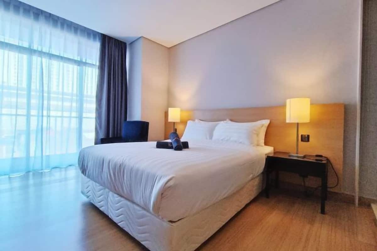 Habitación de hotel con una cama grande, dos lámparas de noche, una silla y vistas a través de cortinas translúcidas.