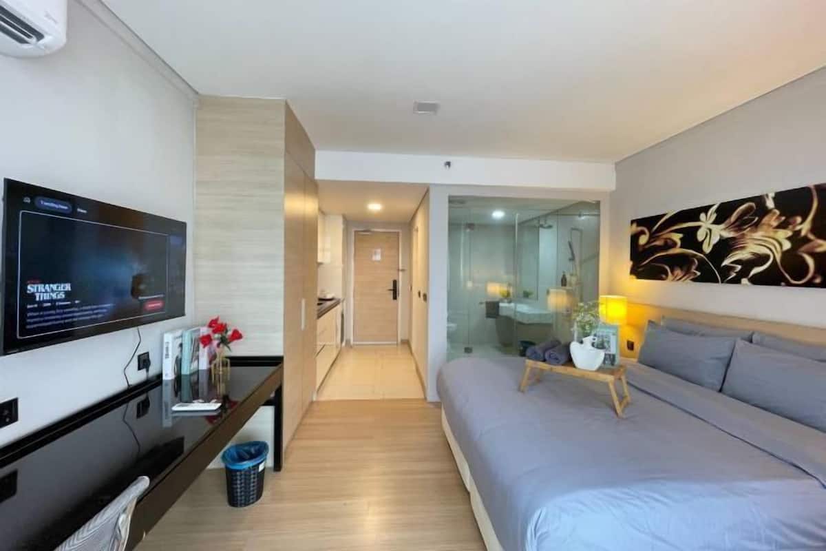 Habitación de hotel moderna con televisión de pantalla plana, sofá, mesita de noche con lámpara y cabina de ducha de vidrio.