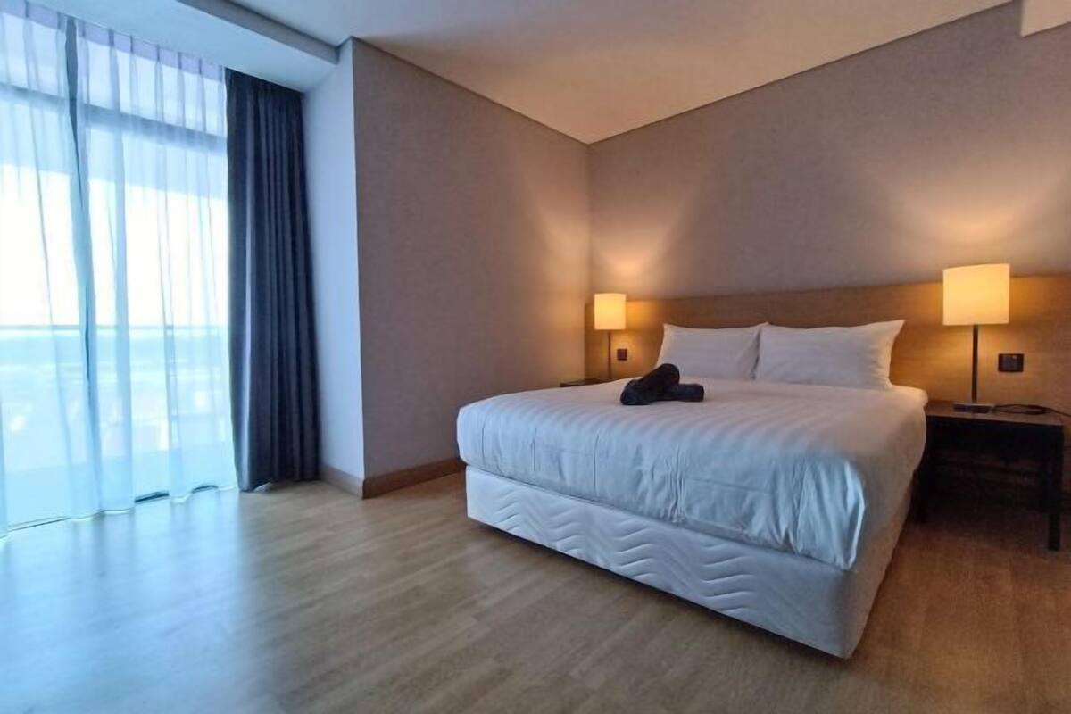 Habitación de hotel con una cama grande, dos lámparas de noche, vistas a través de cortinas traslúcidas y suelo de madera.