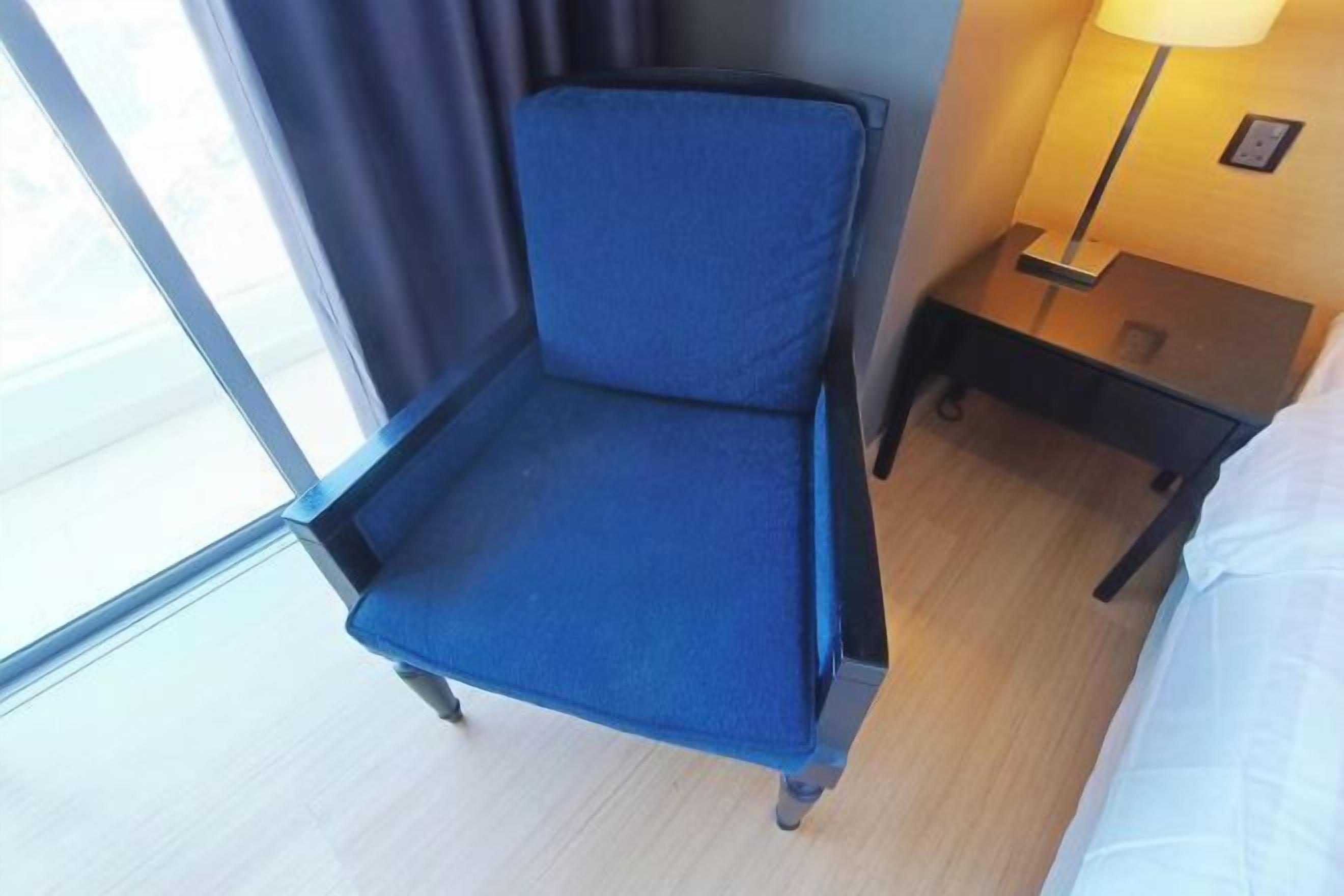 A hotel room with a single blue chair, a bedside table with a lamp, and a bed with white linens.