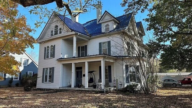 Une maison blanche à deux étages, avec un toit bleu, une véranda avec des chaises et un vélo garé devant.