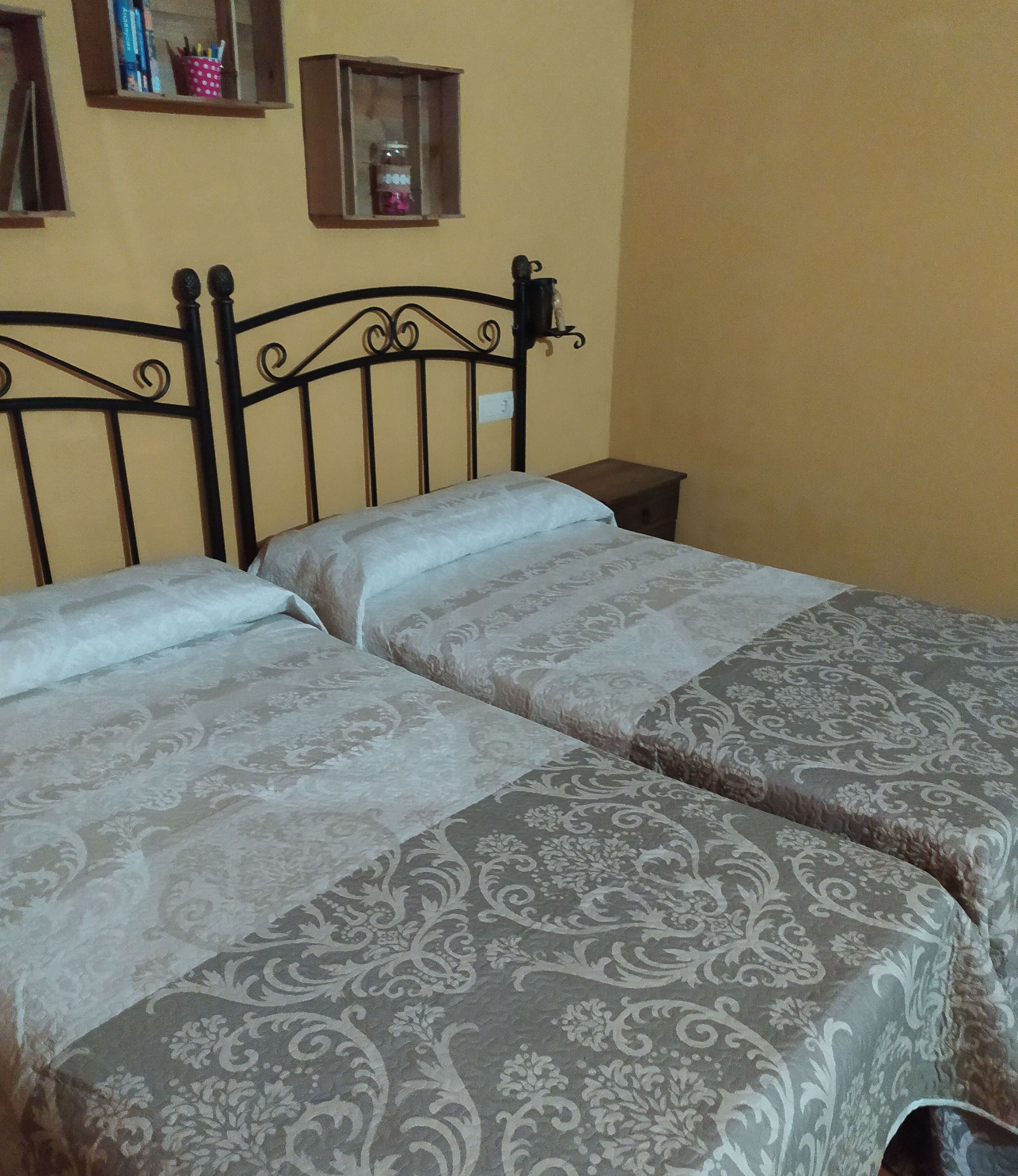 A bed with a metal frame and patterned bedding, a bedside table, and a shelf with books and a jar.