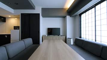 A modern living room with a long wooden table, grey sofas, a flat-screen TV, and a large window providing natural light.