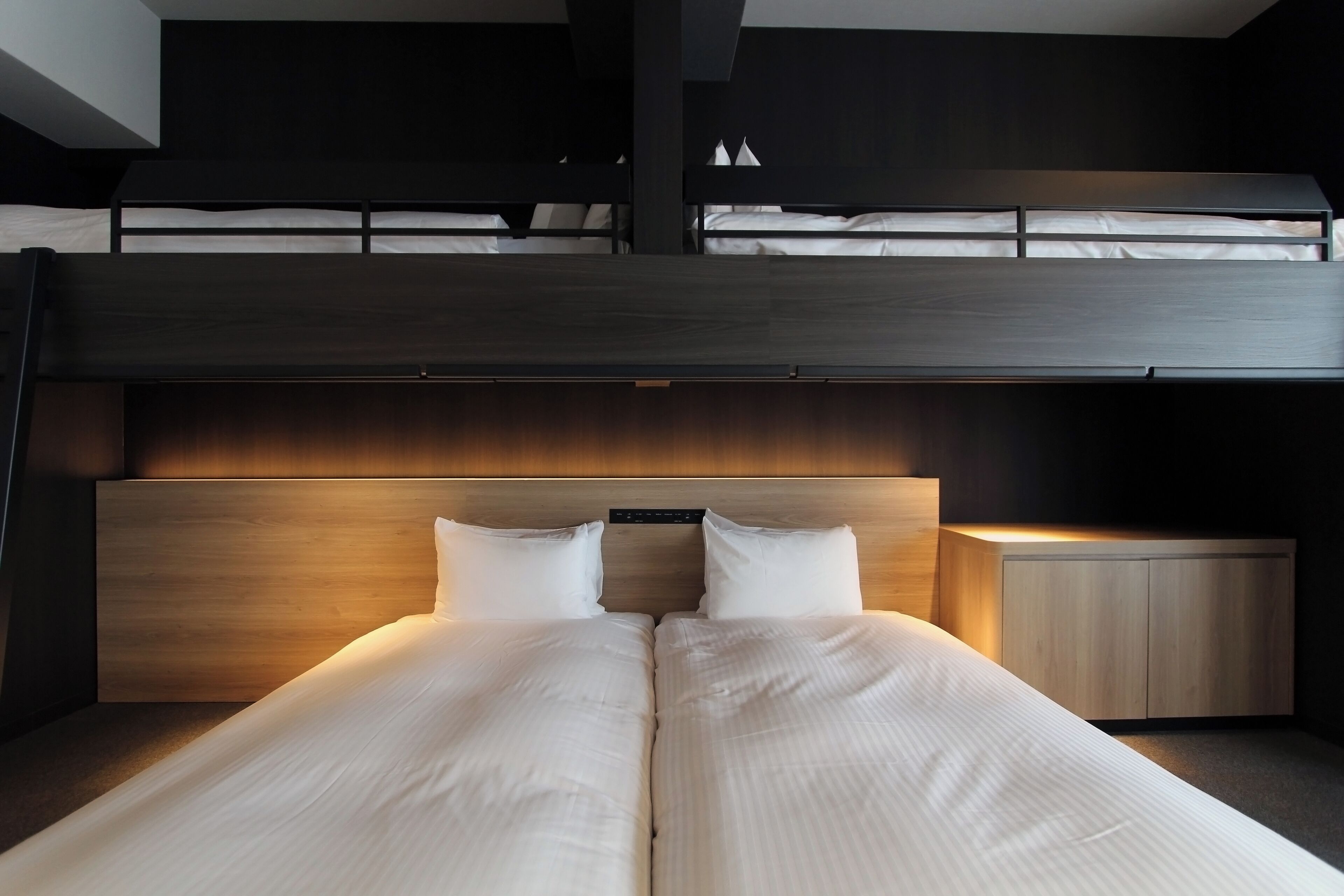 Una habitación de hotel moderna con una litera, un cabecero de madera y una mesita de noche con una lámpara.