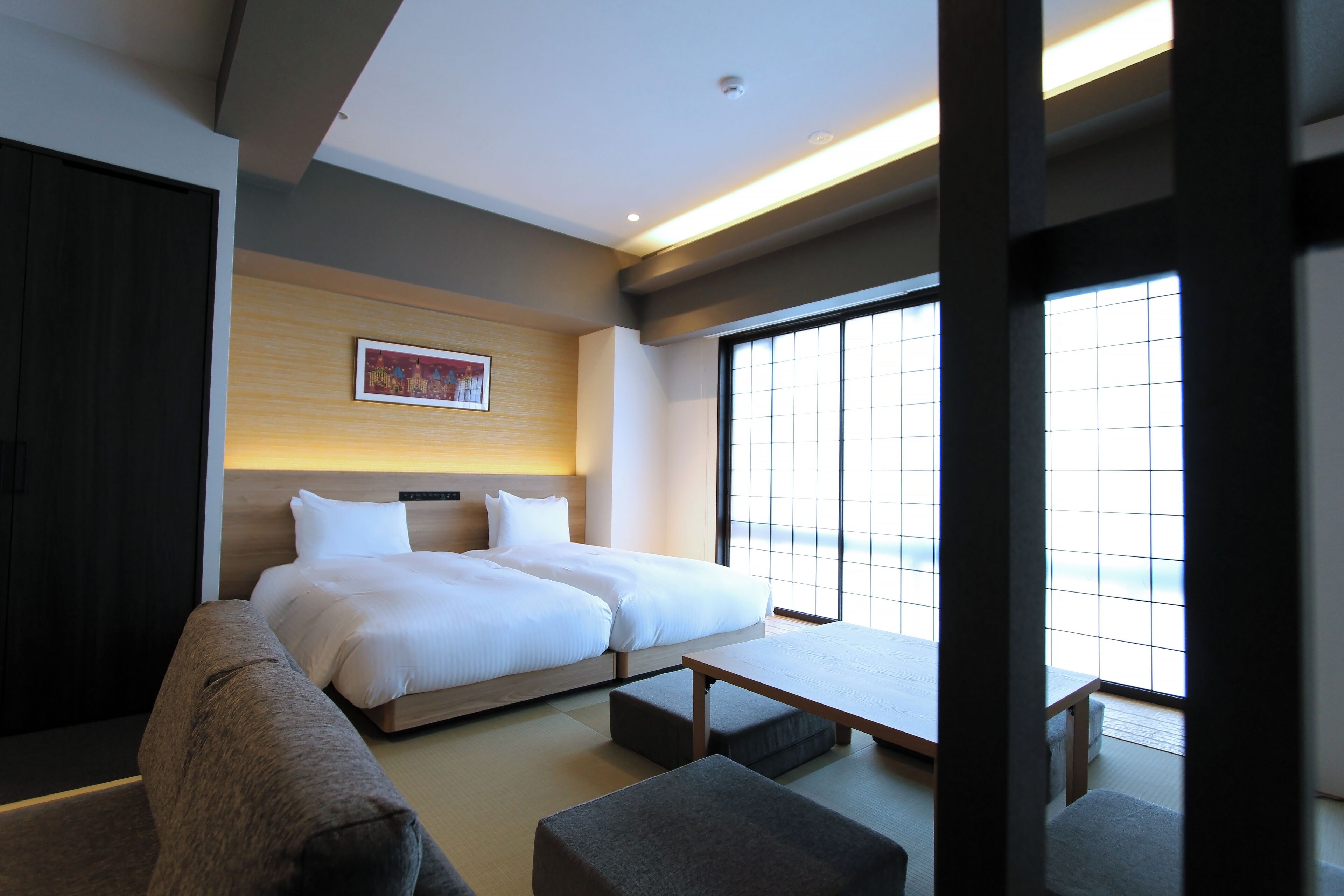 A modern hotel room with a large bed, a sofa, a small table, and a large window providing a view.