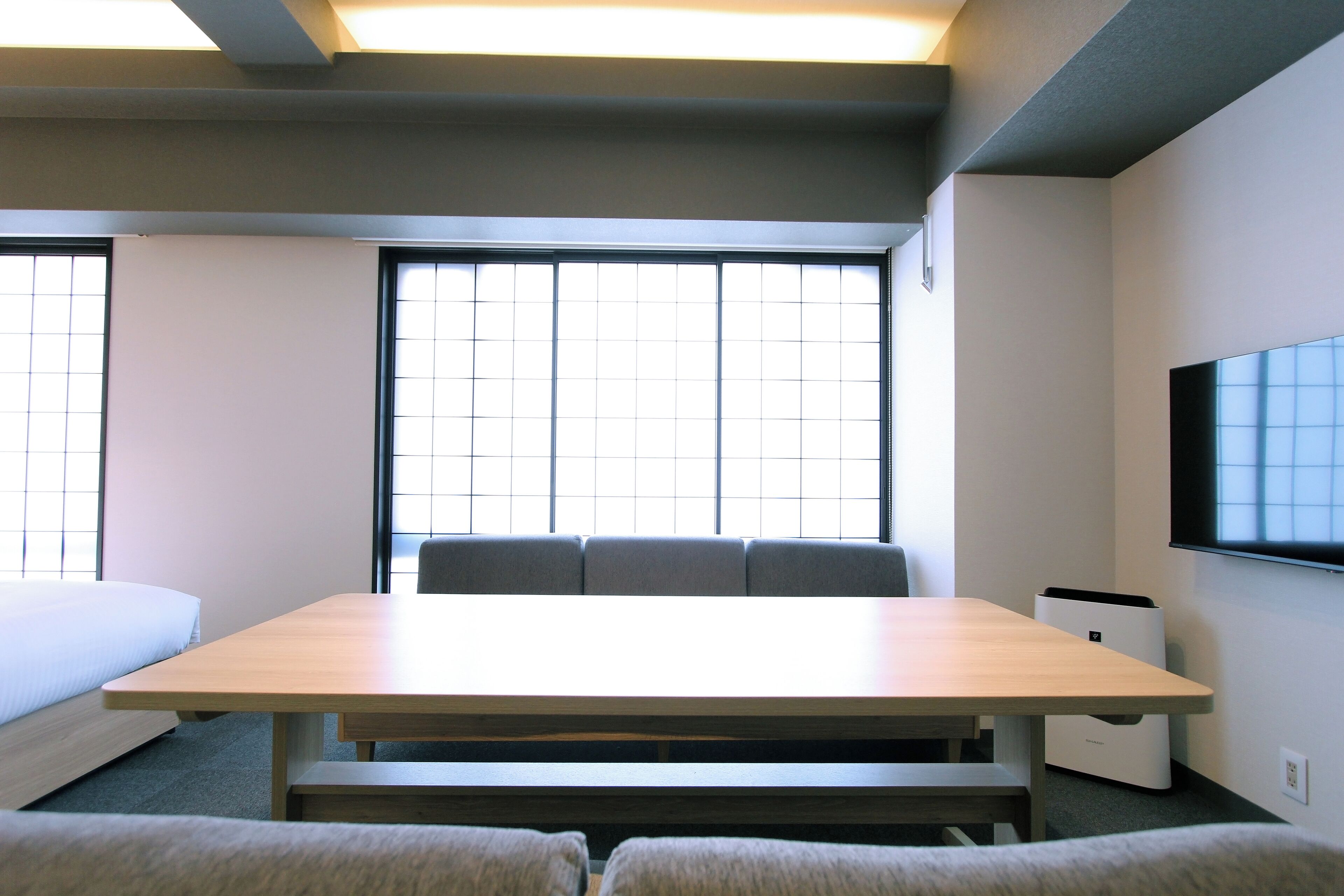 A hotel room with a wooden table, a sofa, a TV mounted on the wall, and a large window with blinds.