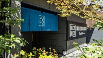 A modern building facade with a blue banner displaying the 'KOKO HOTEL' logo, a gated entrance, and a small outdoor seating area with tables and chairs.