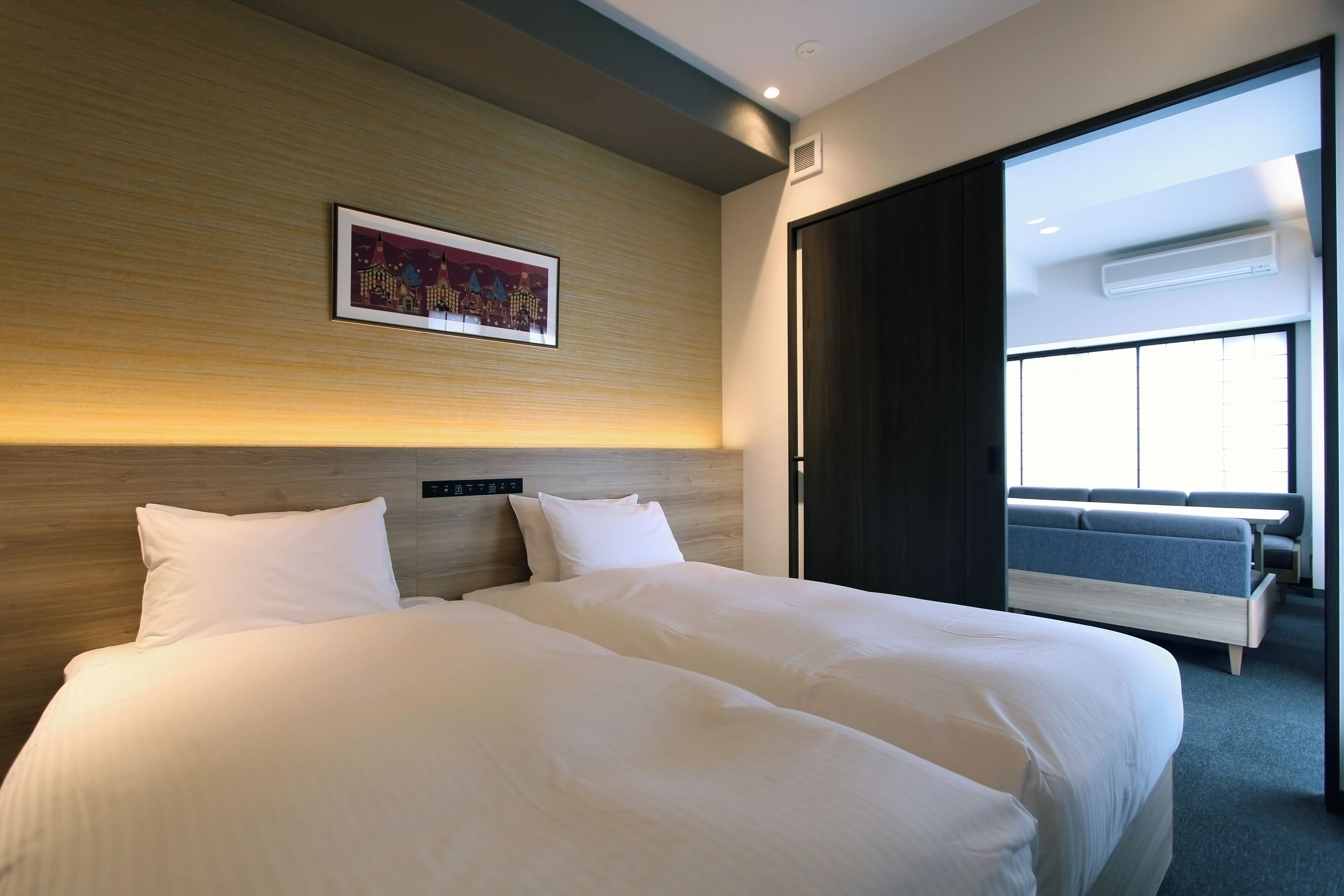 A modern hotel room with a large bed, a headboard with a cityscape artwork, and a view of a balcony with seating.