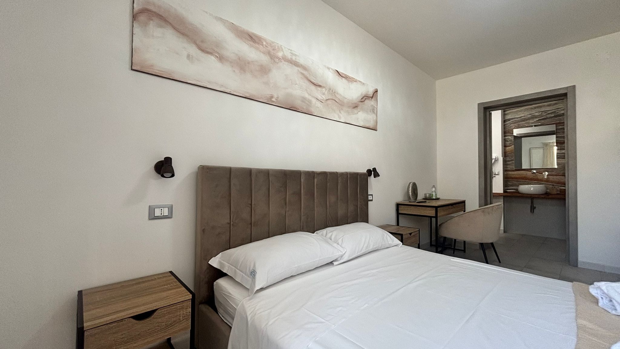 A bedroom with a bed, bedside tables, a desk, a chair, a bathroom with a mirror and sink, and a large abstract painting on the wall.