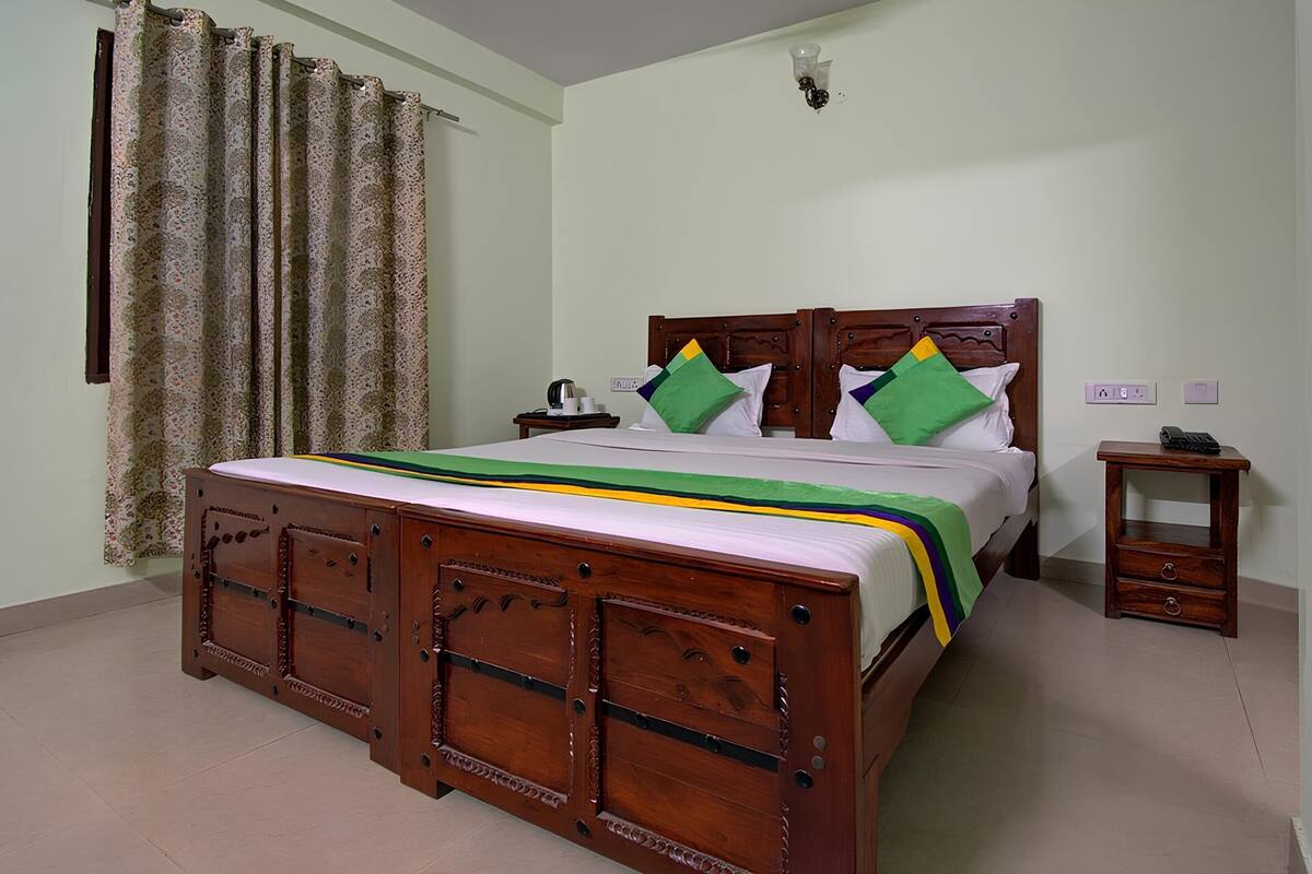 A wooden bed with a green and yellow bedspread, a nightstand with a phone, and a wall-mounted light fixture.
