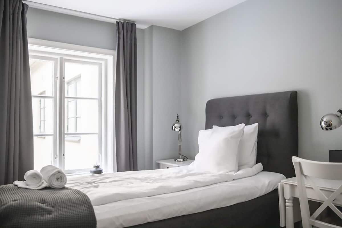 Une chambre d’hôtel avec un lit, un bureau, une chaise, une lampe et une fenêtre avec des rideaux.