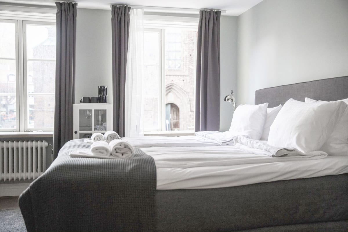 Une chambre d’hôtel avec un grand lit, des rideaux gris, un radiateur et une vue sur un immeuble par la fenêtre.