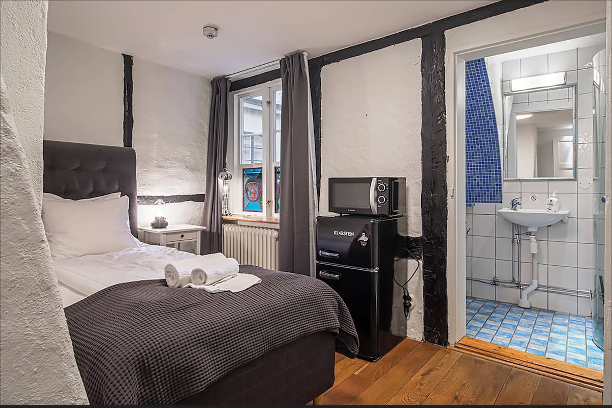 Une chambre à coucher avec un lit, une table de chevet, une fenêtre avec des rideaux, un petit appareil électroménager noir et une salle de bain avec un lavabo et un sol carrelé.