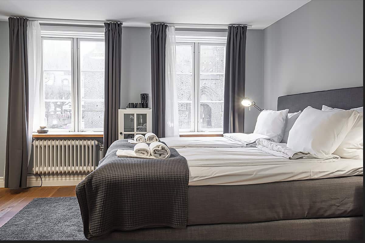 Une chambre à coucher avec un grand lit, une table de chevet, une lampe et une vue sur le paysage urbain par la fenêtre.
